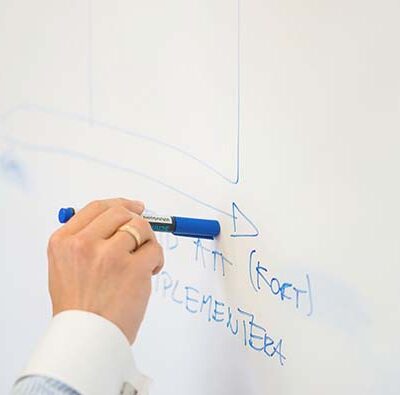 Närbild på en hand som håller i en penna och pekar på en matris på en vit whiteboard tavla.