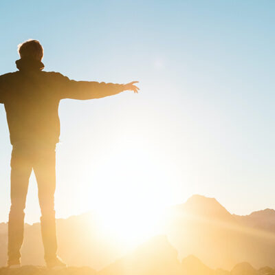 En person står med armarna utsträckta i sidan och tittar mot solen, motljuset gör att vi bara ser hens silhuett.