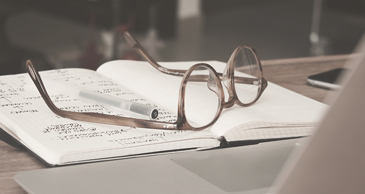 Närbild på ett par glasögon som ligger på en öppen anteckningsbok på ett bord.