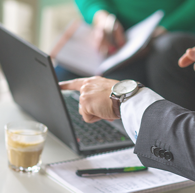 Närbild på en man i kostym som pekar på en laptop, i bakrunden ser man en perosn i grön tröja som sitter med i mötet.