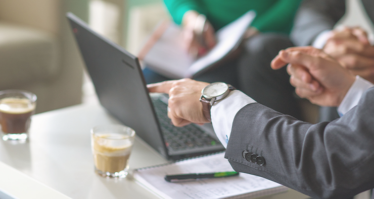 Närbild på en man i kostym som pekar på en laptop, i bakrunden ser man en perosn i grön tröja som sitter med i mötet.